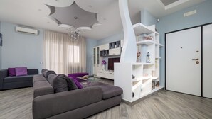 Un salon moderne avec un canapé d’angle gris, une étagère blanche et une grande fenêtre ornée de rideaux légers.