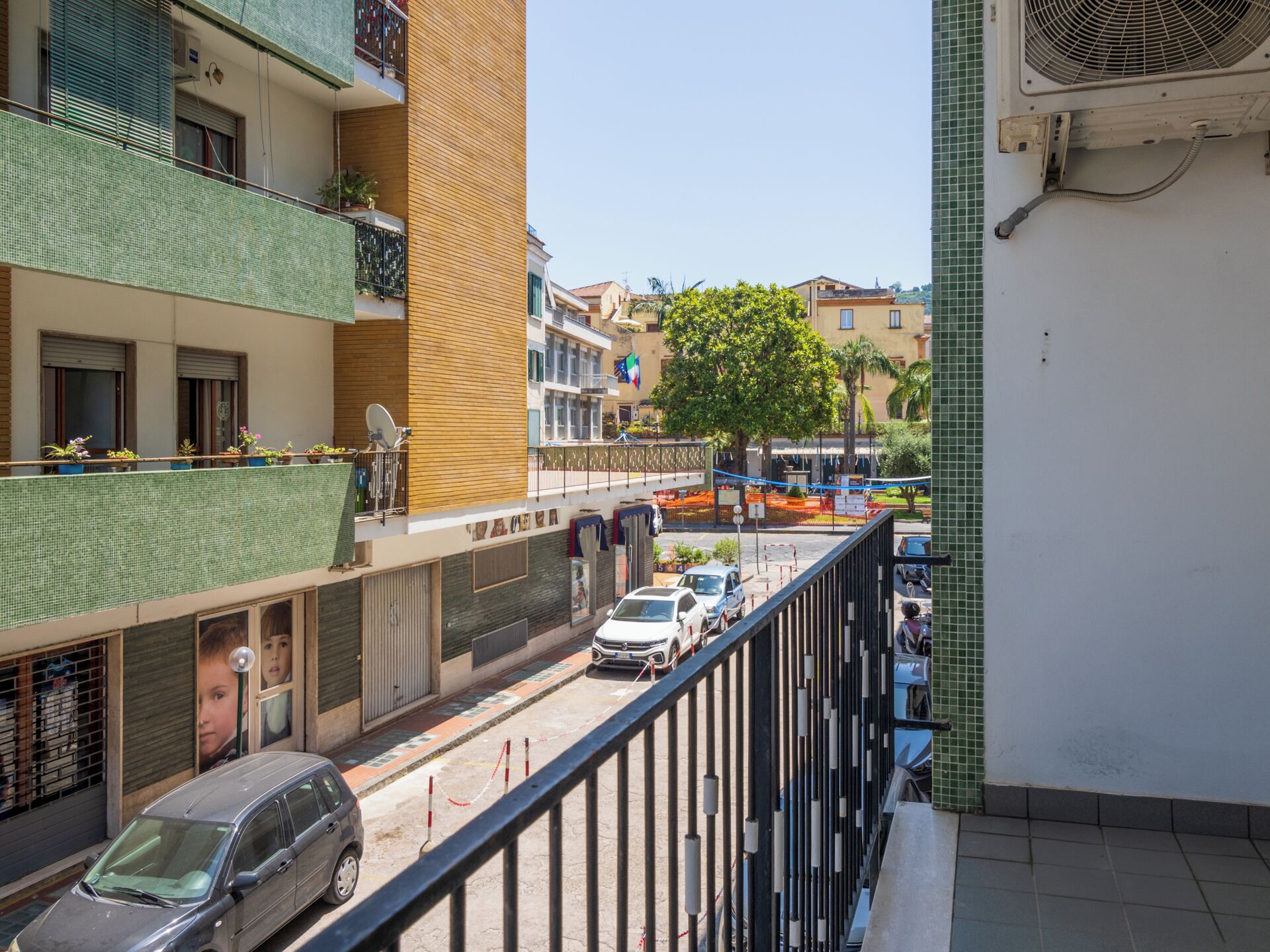 Uma rua residencial com uma fileira de carros estacionados, uma varanda com grade de metal e um edifício com fachada revestida de azulejos.