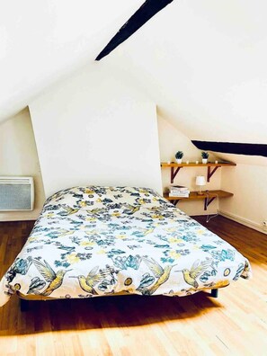 A bedroom with a sloped ceiling, a bed with a floral-patterned bedspread, a small bedside table with a lamp, and a shelf with potted plants.