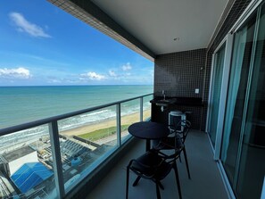 A balcony with a view of the ocean and beach, a small table and chairs, and a tiled backsplash.