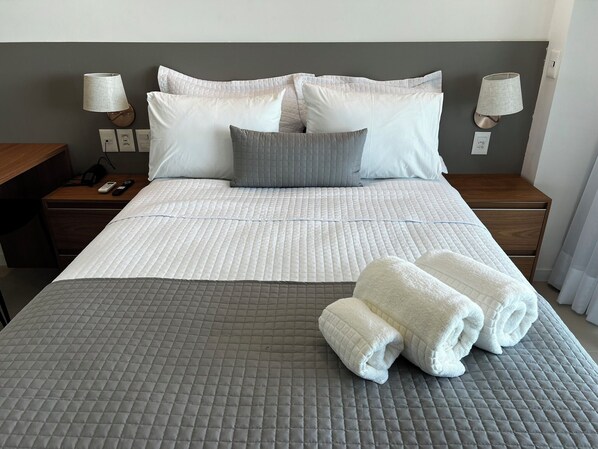 A neatly made bed with a grey quilt, white pillows, and a grey throw blanket. There are two bedside lamps, a wooden nightstand, and a folded towel on the bed.
