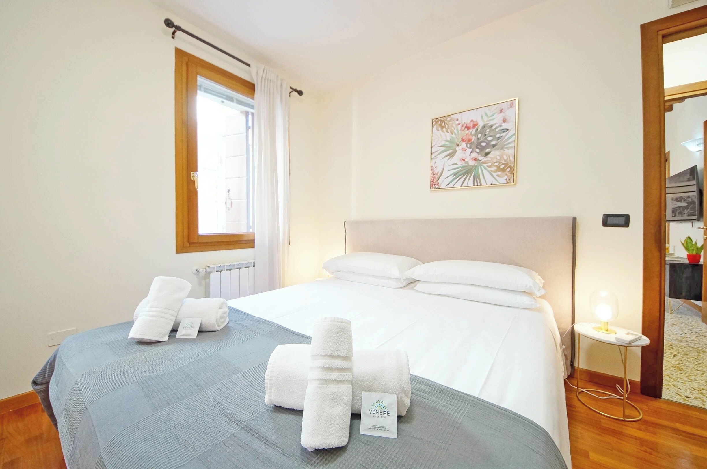 A hotel room with a bed, a window with curtains, a bedside table with a lamp, and a framed picture on the wall.