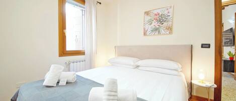 A hotel room with a bed, a window with curtains, a bedside table with a lamp, and a framed picture on the wall.
