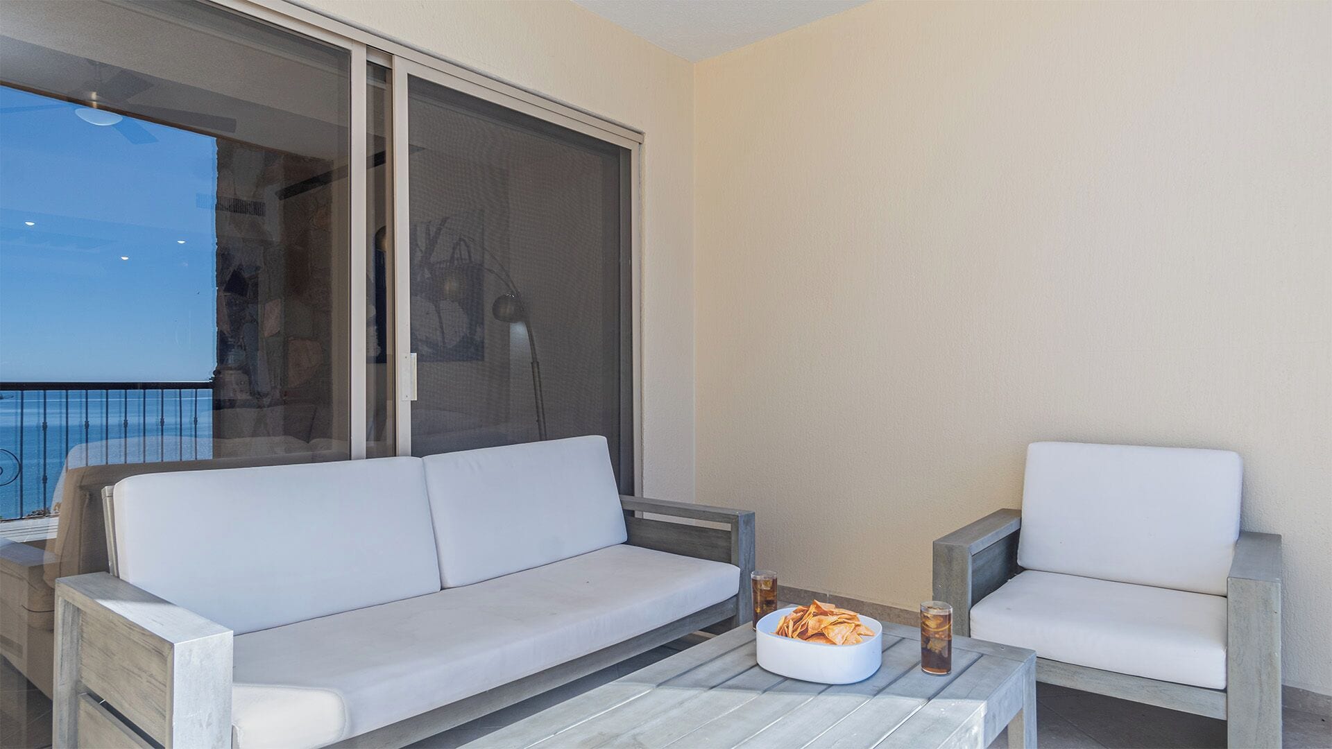 Ein Balkon mit einer weißen Couch und einem Sessel, ein kleiner Tisch mit Snacks und Getränken und Blick aufs Meer.