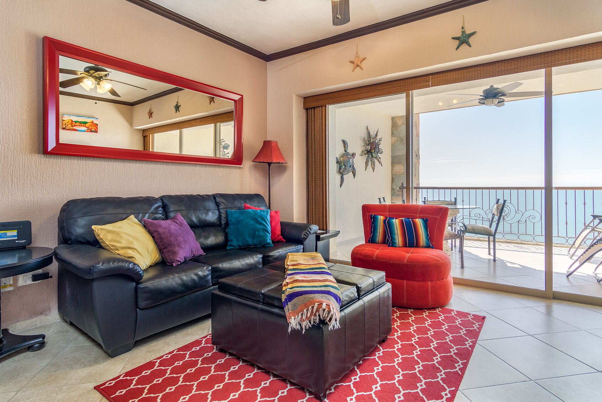 Un salón con un sofá de cuero negro, un sillón rojo y una mesa de centro con una manta a rayas. Se observa un espejo grande en la pared, una lámpara de pie roja y vistas al océano a través de una puerta corrediza de vidrio.