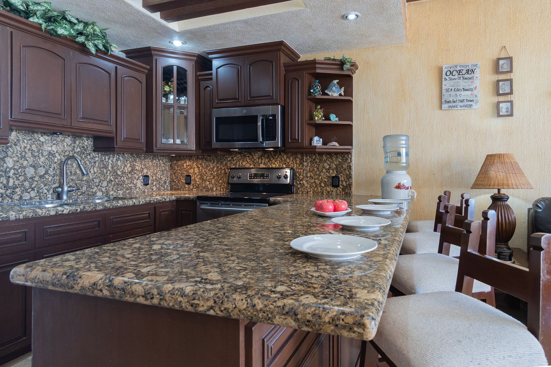 A kitchen with granite countertops, dark wooden cabinets, a microwave, stove, and a water dispenser. There are plates and apples on the counter.