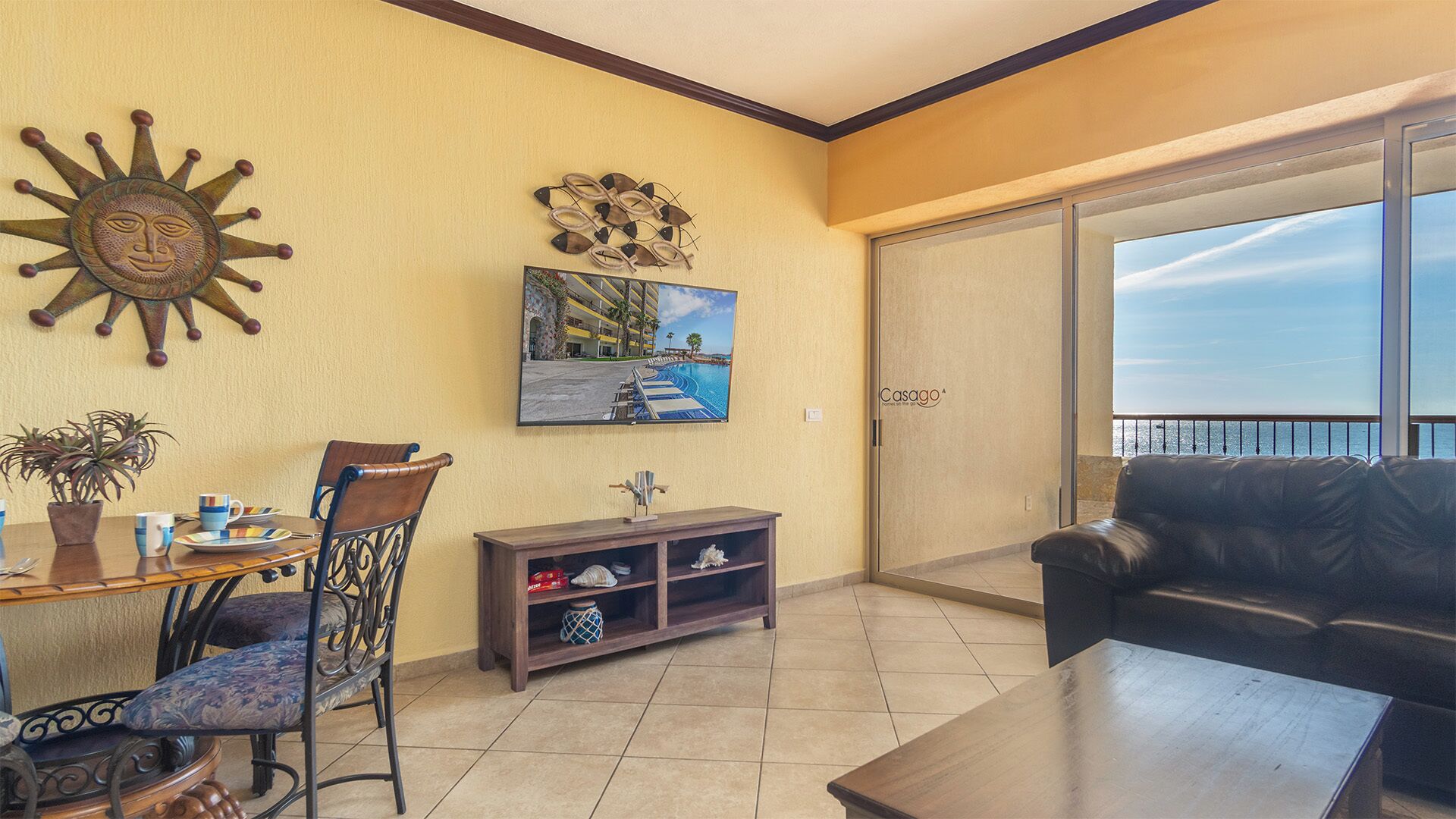 A living room with a sofa, a dining table with chairs, a TV mounted on the wall, and a balcony with a view.