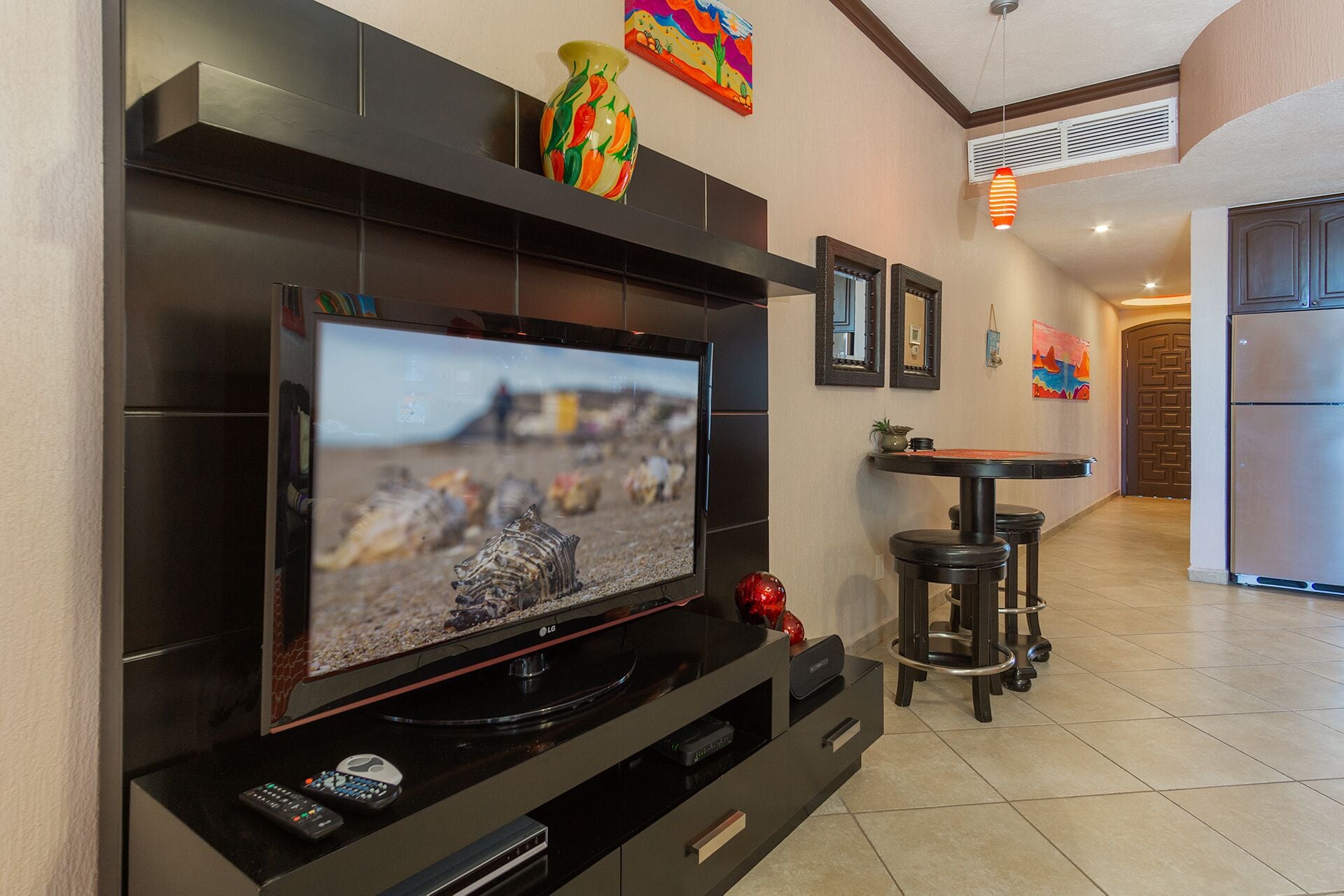 A modern living room with a flat-screen TV mounted on a black entertainment unit, a small round table with stools, and a kitchen area visible in the background.