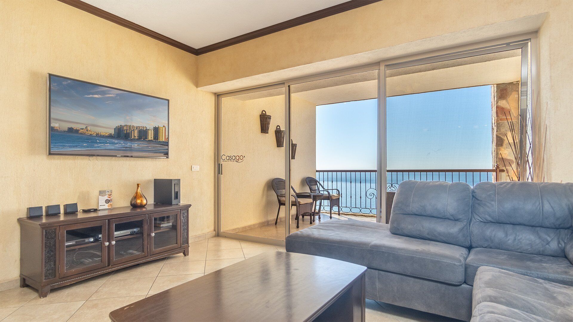 Un soggiorno con divano, tavolino, televisione e mobile. È presente un balcone con vista sul mare.