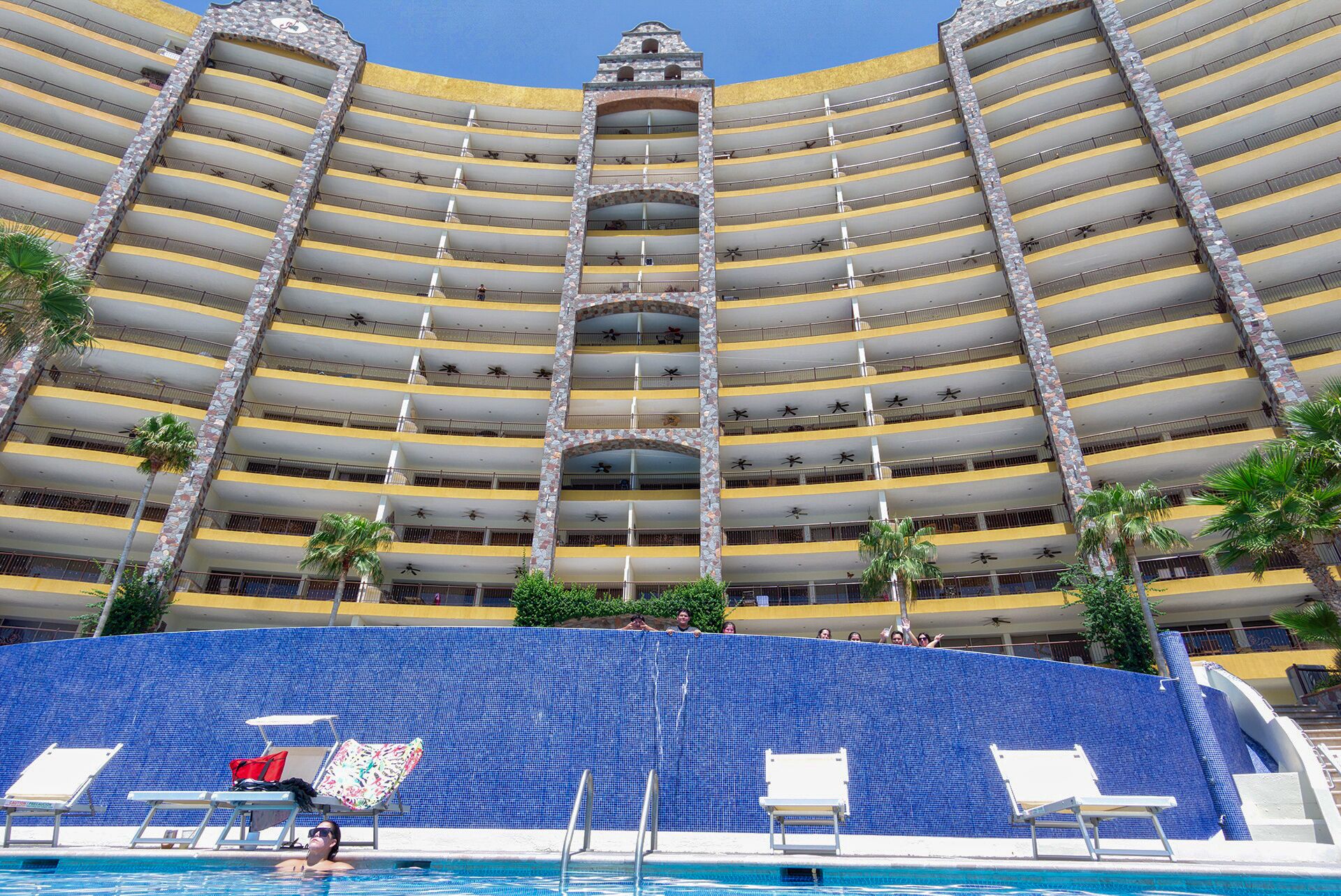 Un hotel a più piani con area piscina, caratterizzato da un muro in piastrelle blu, numerosi balconi e palme.