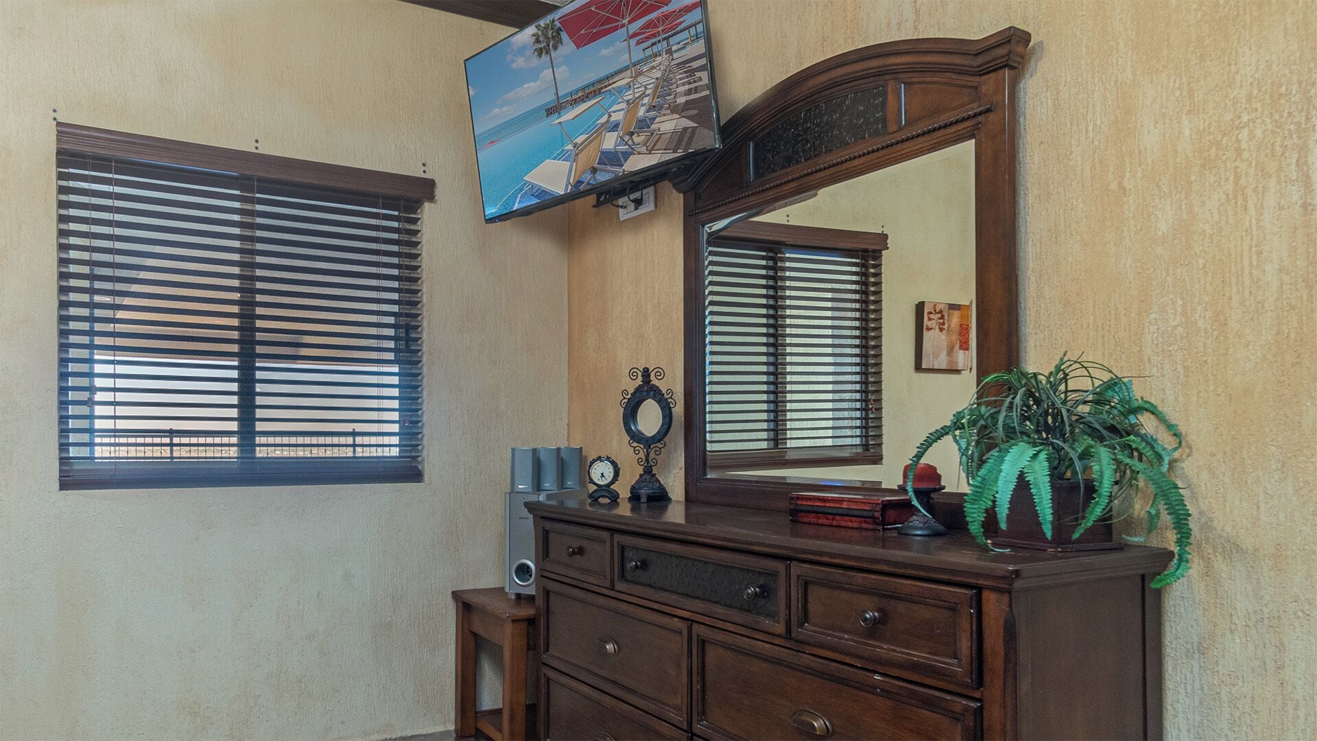 A room with a wooden dresser, a large mirror, and a television mounted on the wall.