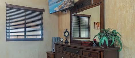 A room with a wooden dresser, a large mirror, and a television mounted on the wall.