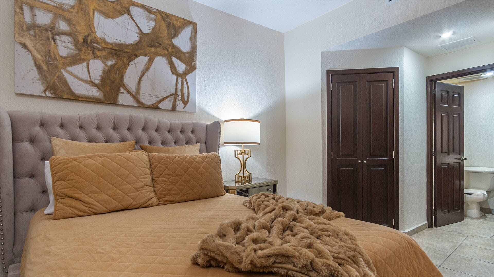 A bedroom with a tufted headboard, a large abstract painting, a bedside lamp, and a fluffy throw blanket.