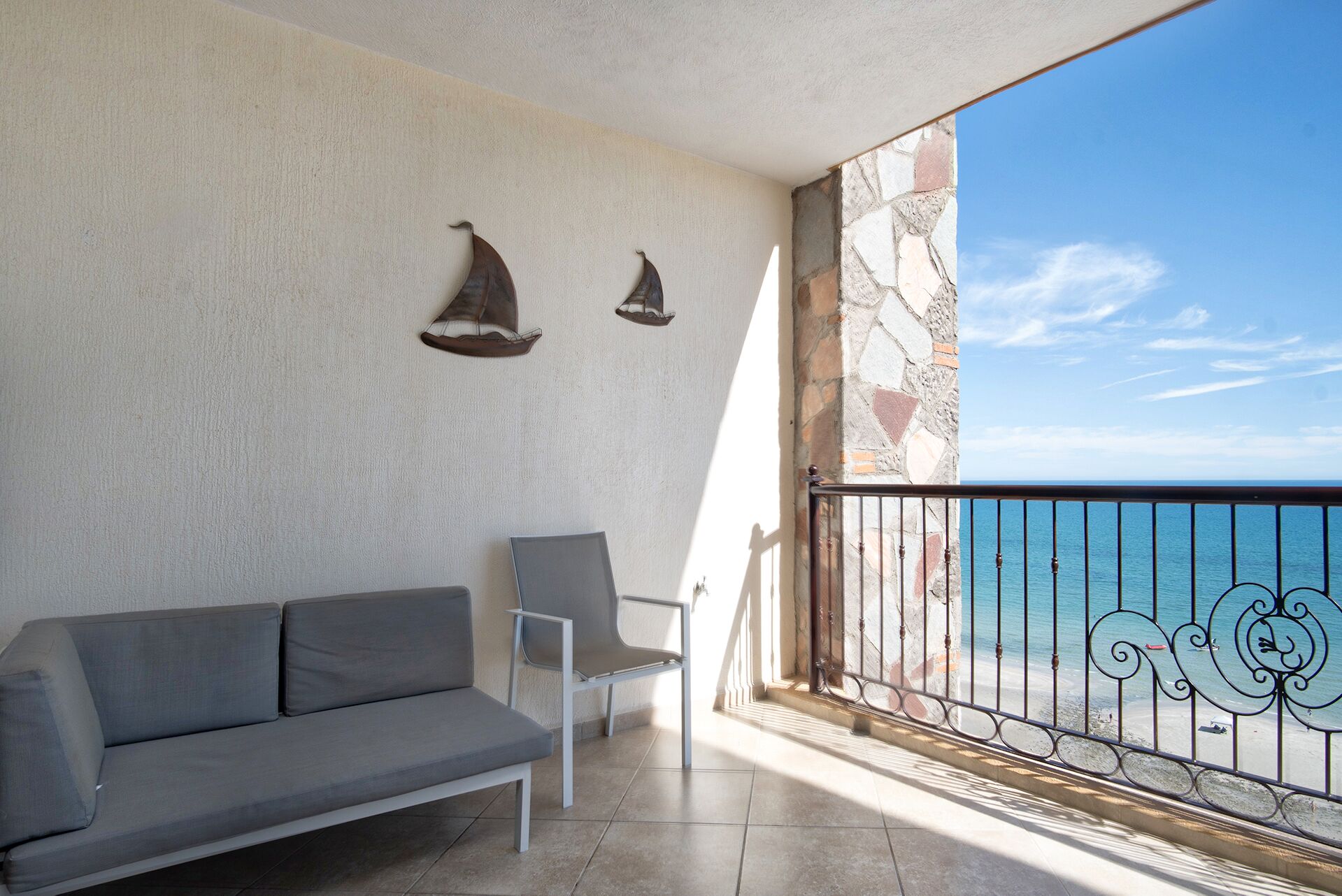 Un balcone con un divano grigio, una poltrona bianca, un muro in pietra e vista sul mare.