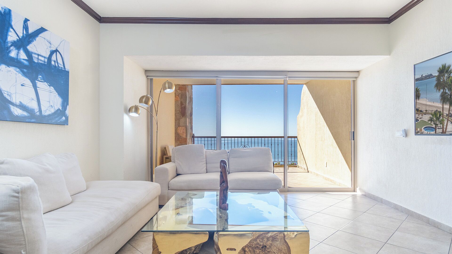 A modern living room with a glass coffee table, a white sofa, and a view of the sea through a sliding glass door.