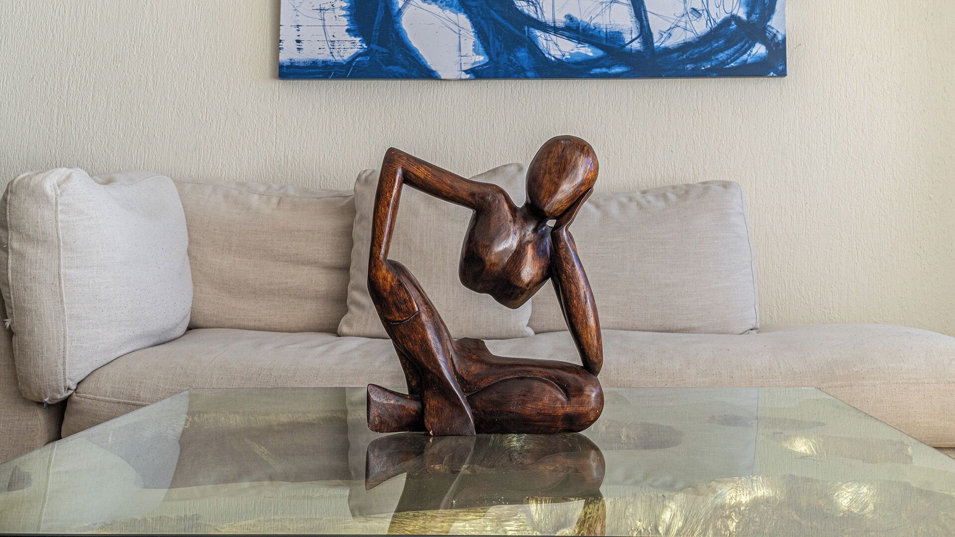 A sculpture on a glass table in front of a beige sofa with a blue abstract painting on the wall.