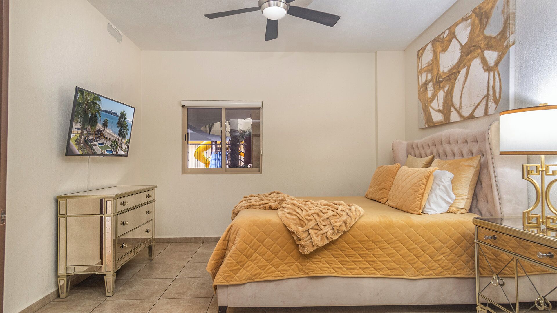 Un dormitorio con cama, mesita de noche, cómoda, ventilador de techo, ventana y un cuadro en la pared.