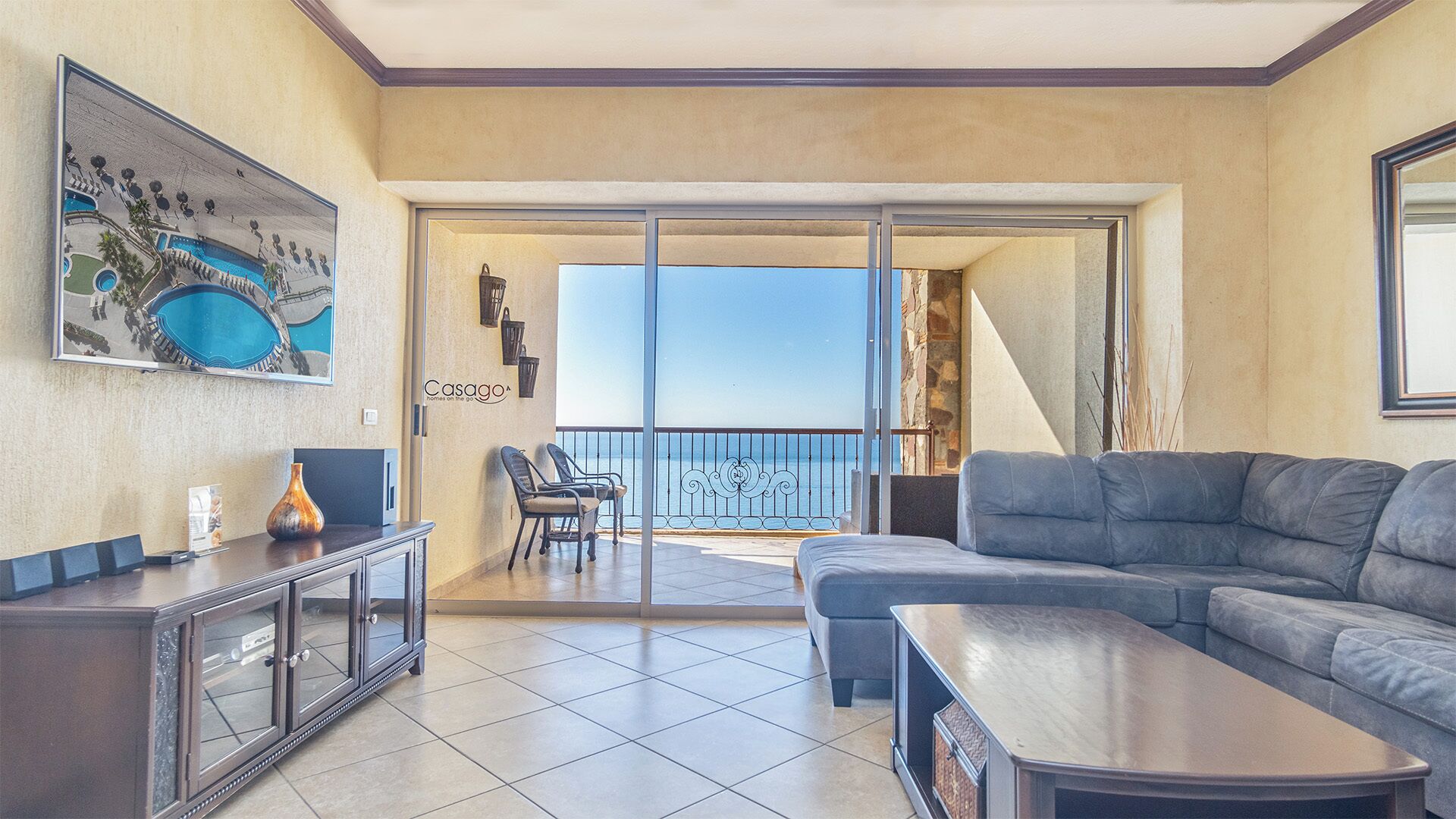 Una sala de estar moderna con un sofá gris, una mesita de centro y un televisor sobre un mueble. Cuenta con un balcón con vista al mar y un cuadro en la pared.