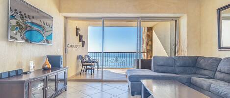 Una sala de estar moderna con un sofá gris, una mesita de centro y un televisor sobre un mueble. Cuenta con un balcón con vista al mar y un cuadro en la pared.