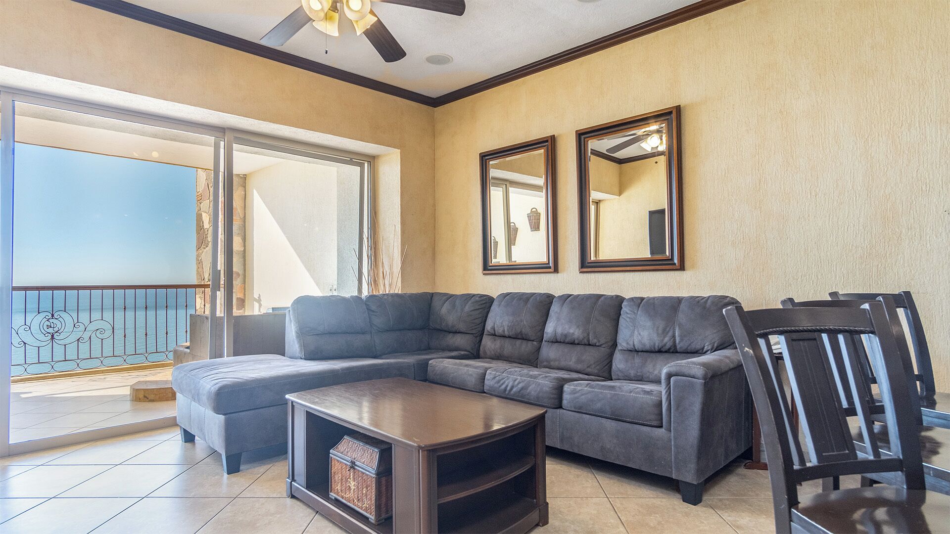 A living room with a grey sectional sofa, a small wooden coffee table, and a black chair. There are two framed mirrors on the wall, a ceiling fan, and a balcony with a railing.