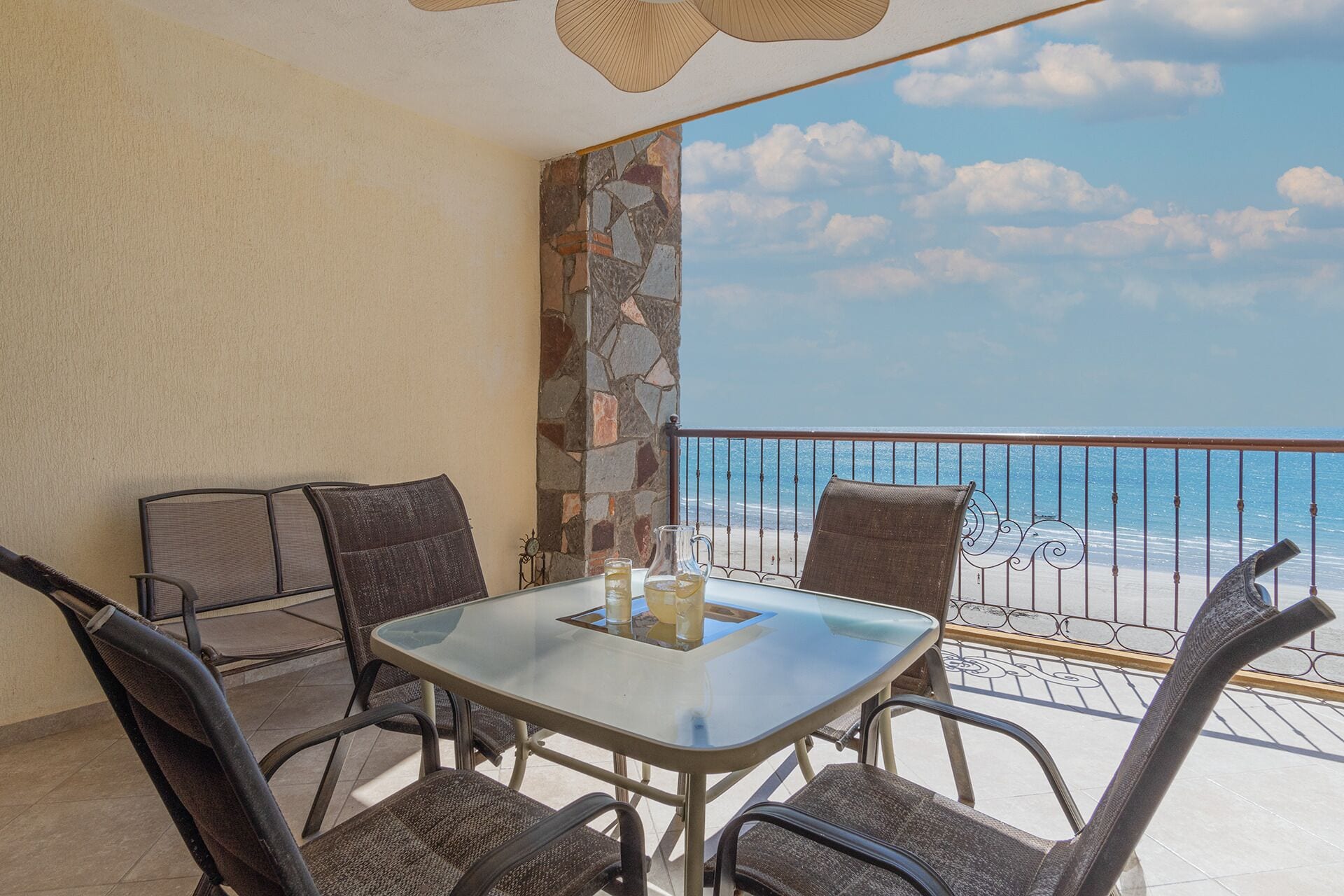 A balcony with a glass table, four chairs, and a view of the beach and ocean.