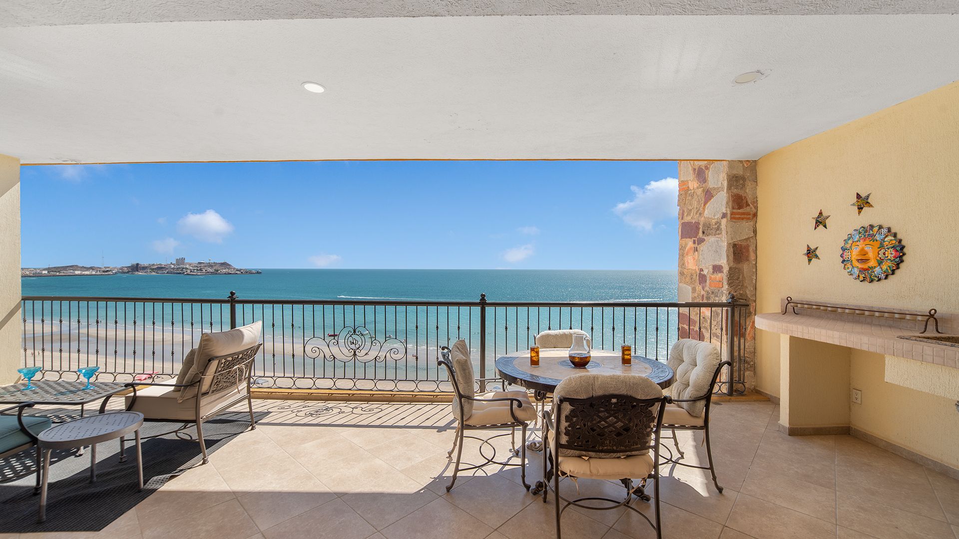 A balcony with a view of the sea, a table with chairs, and a decorative wall piece.