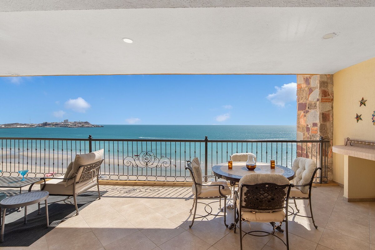 Un balcón con vista al mar, una mesa con sillas y un adorno en la pared.