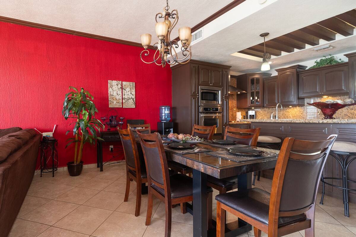 Un comedor con pared roja, mesa de madera y sillas. Hay una araña colgando sobre la mesa.