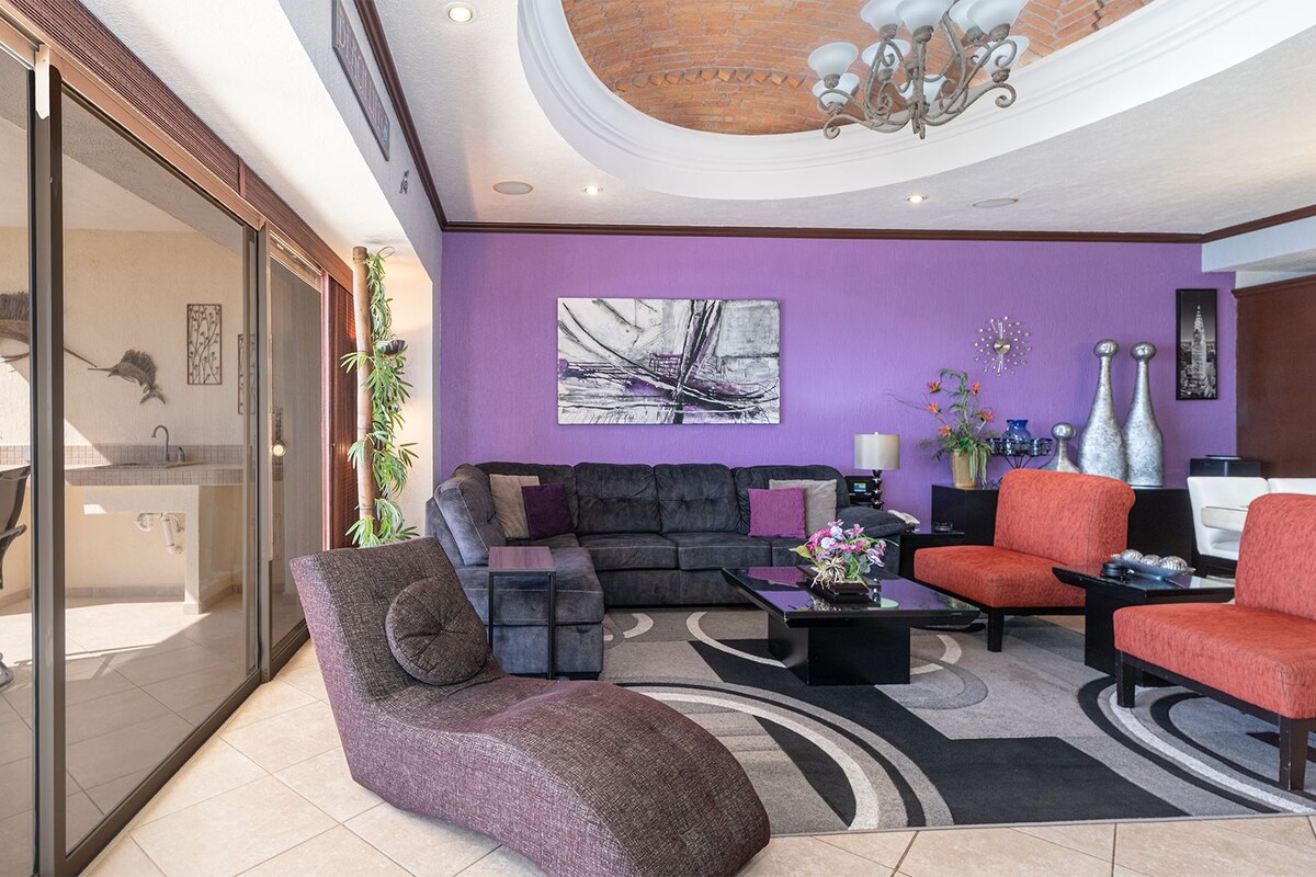 A modern living room with a purple wall, a black sofa, a red armchair, and a glass-enclosed patio area with a view of the outside.