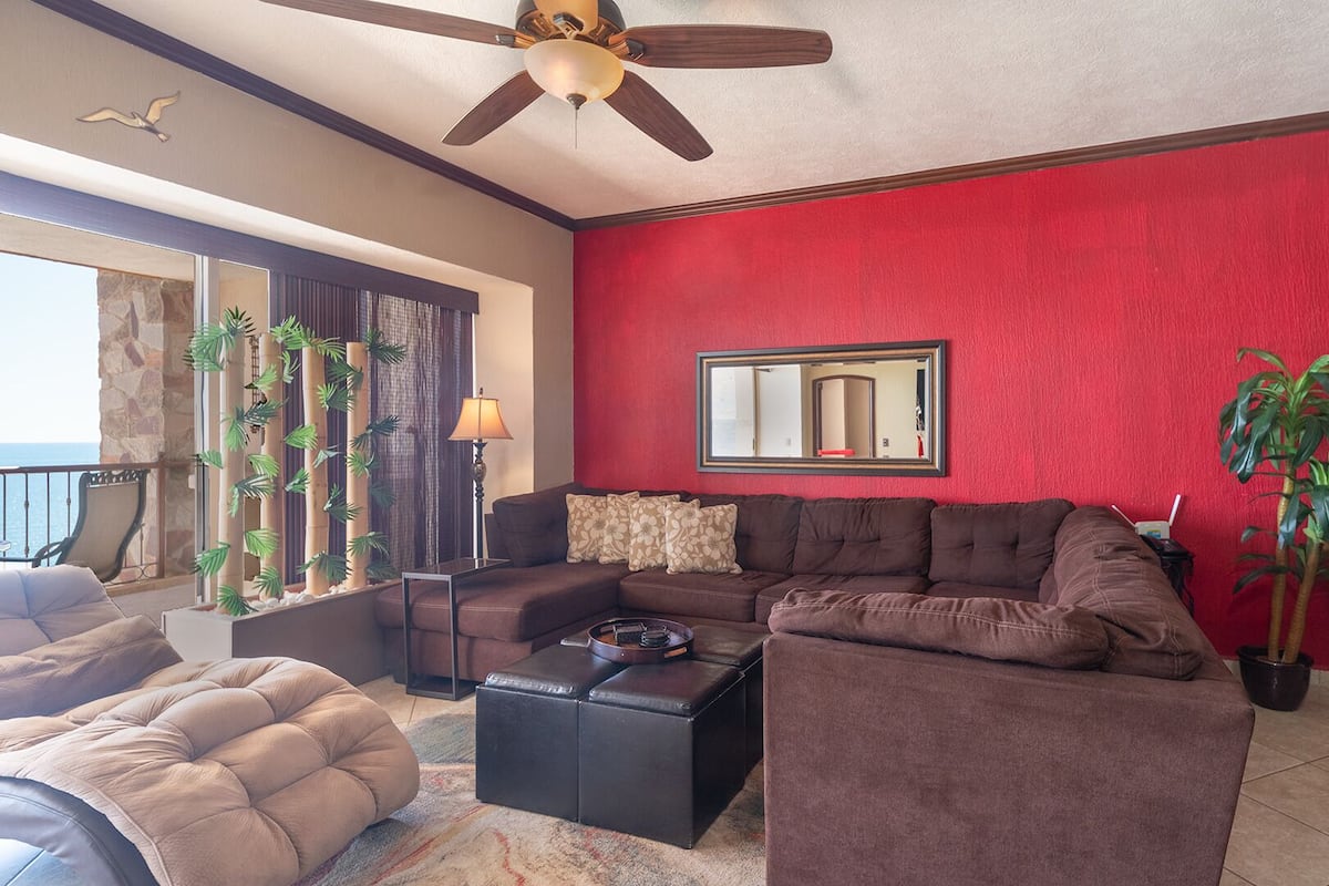 Un salon avec un mur rouge, un ventilateur de plafond, une grande fenêtre et une vue sur l’océan.