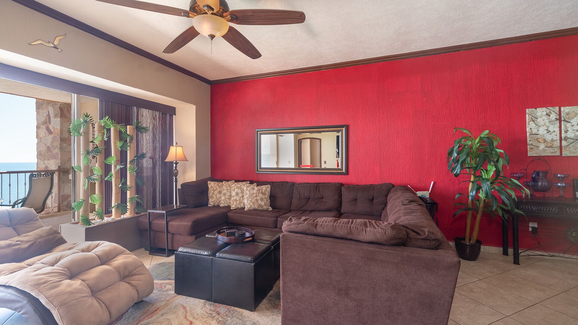 Un salon avec un mur rouge, un ventilateur de plafond, une grande fenêtre et une vue sur l’océan.