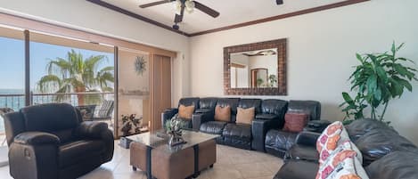 Un salon avec un canapé, un fauteuil et une table basse. On y trouve également un ventilateur de plafond, une grande fenêtre et une plante.