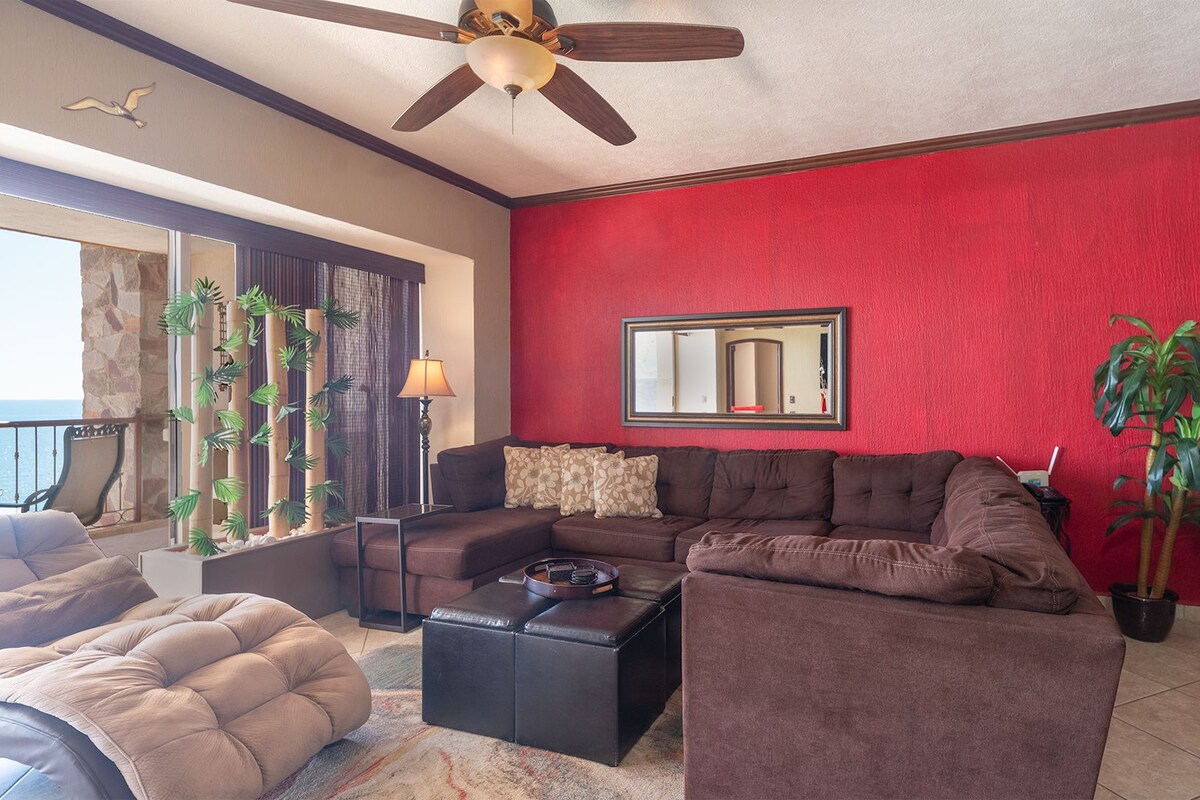 A living room with a red wall, a ceiling fan, a large window, and a view of the ocean.