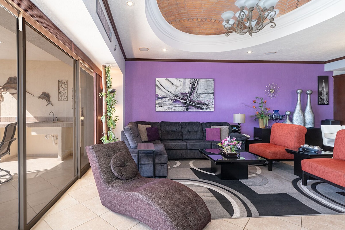 Una sala de estar moderna con una pared morada, un sofá negro, un sillón rojo y un área de patio con vidrio que ofrece vistas al exterior.