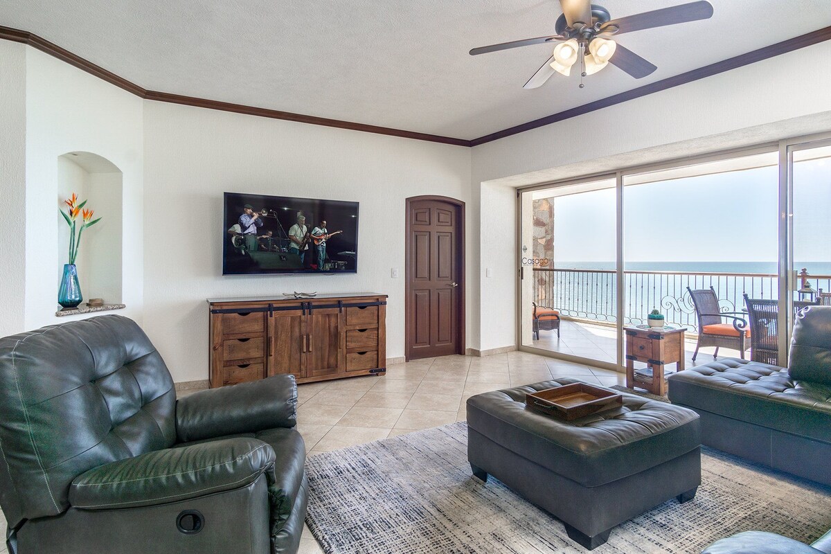 A living room with a television, leather furniture, a wooden cabinet, and a balcony with a view.