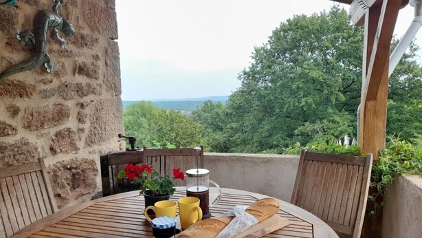 Et rundt træbord med et baguette, en kaffepotte og to kopper på en balkon med udsigt over et skovlandskab.