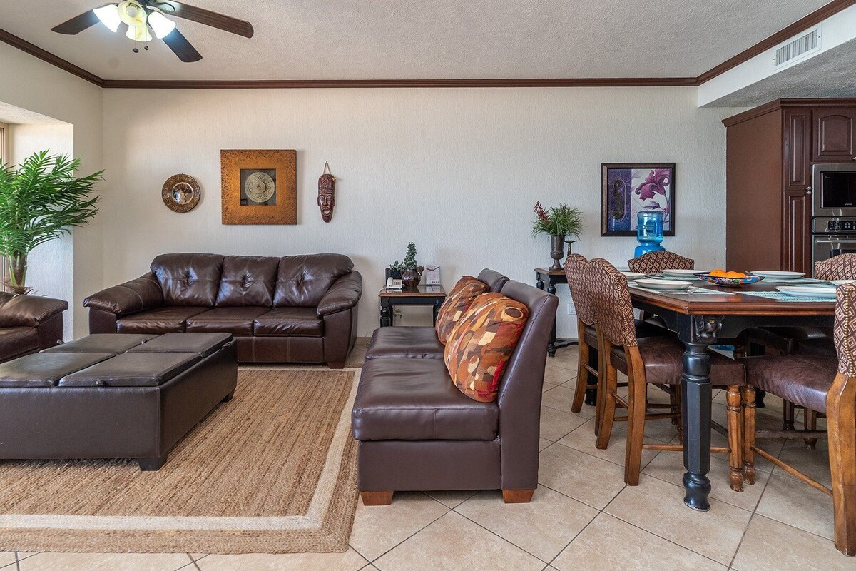 Un vaste salon doté d’un canapé en cuir marron, d’un pouf et d’un fauteuil assorti. Un coin repas avec une table et des chaises en bois est situé à côté.