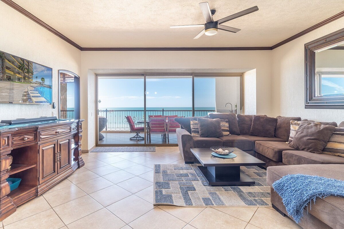 A spacious living room with a balcony, a wooden desk, and a large window offering a view of the ocean.