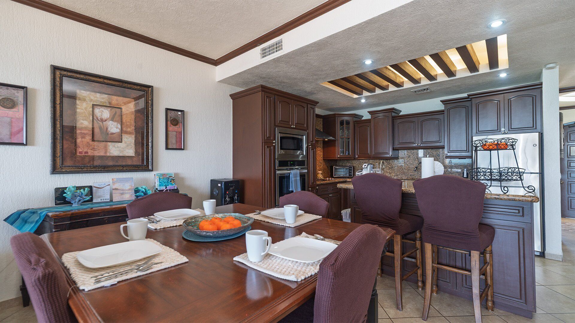 Uma sala de jantar com uma mesa de madeira posta para quatro pessoas, rodeada por cadeiras de madeira escura. Há quadros nas paredes, e cozinha com armários de madeira escura e eletrodomésticos de aço inoxidável pode ser vista ao fundo.