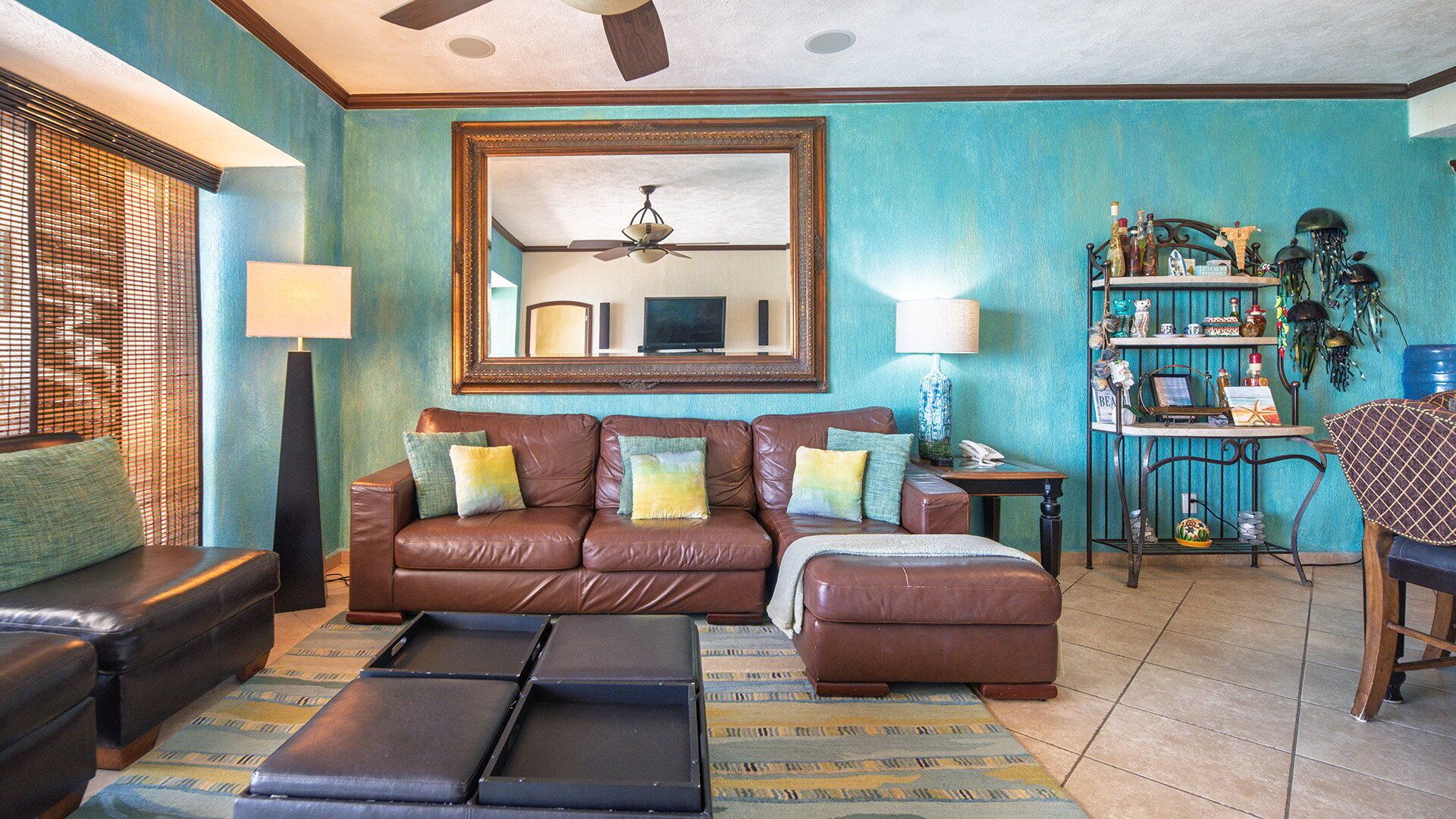 A living room with a brown leather sofa, a coffee table, and a TV. There is a ceiling fan, a lamp, and a shelf with various items.