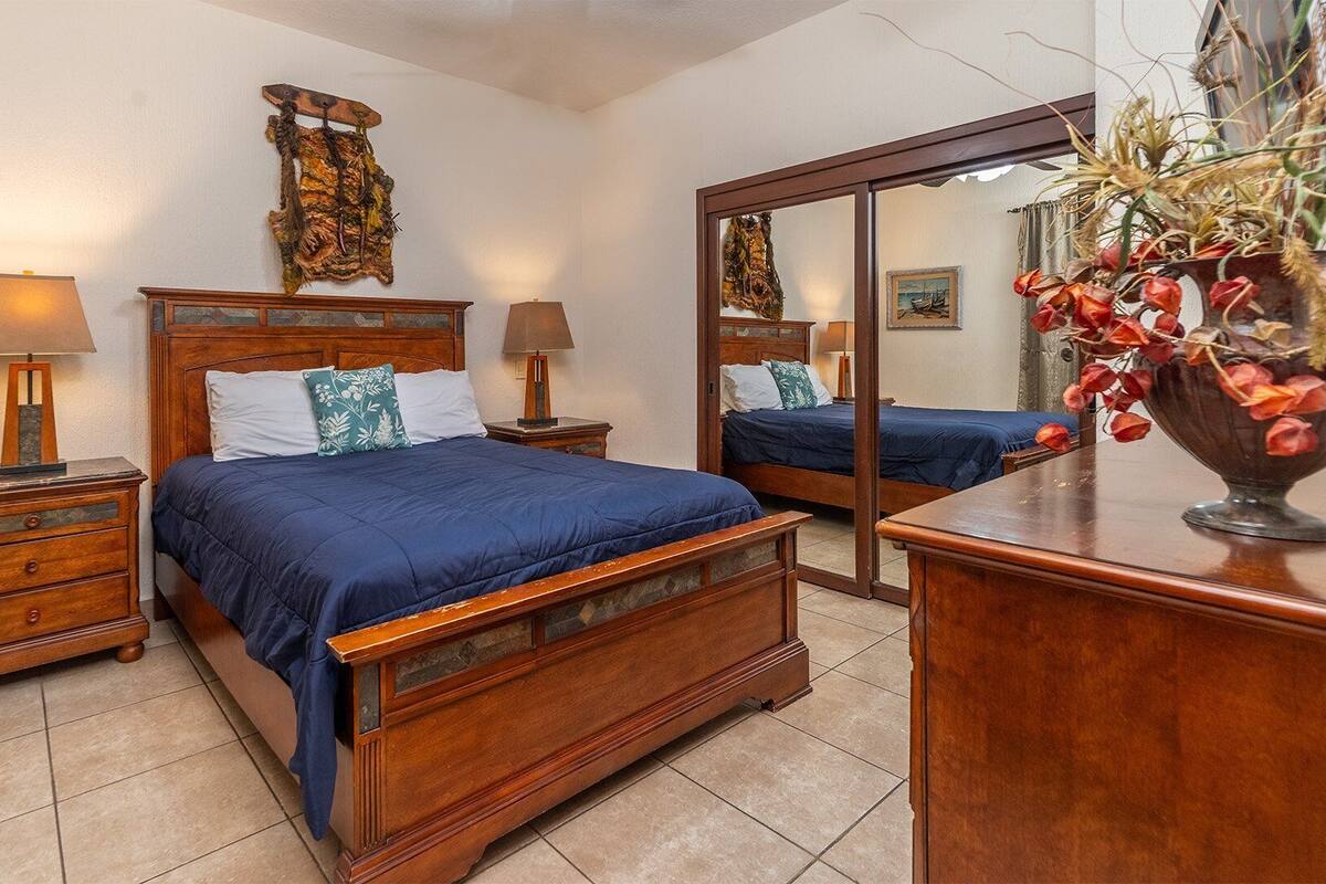 Een slaapkamer met een houten bed, twee nachtkastjes, een dressoir, een spiegel en een vaas met bloemen.
