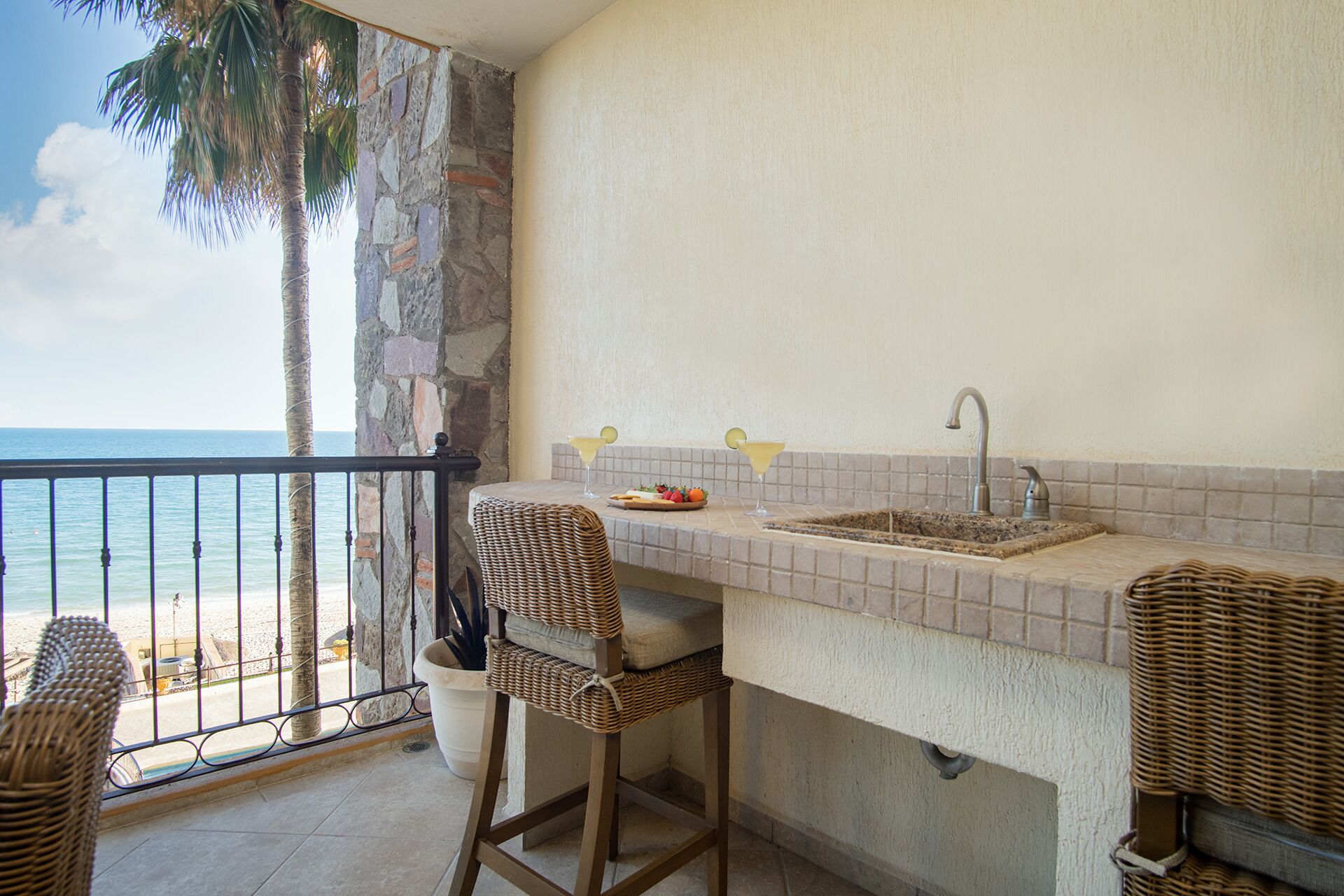 A balcony with a view of the sea, a small bar area with a sink and stools, and wicker furniture.