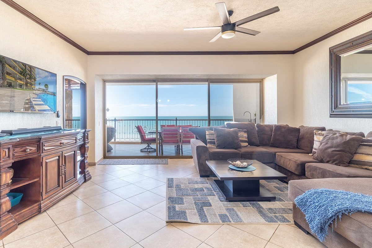 A spacious living room with a balcony, a wooden desk, and a large window offering a view of the ocean.