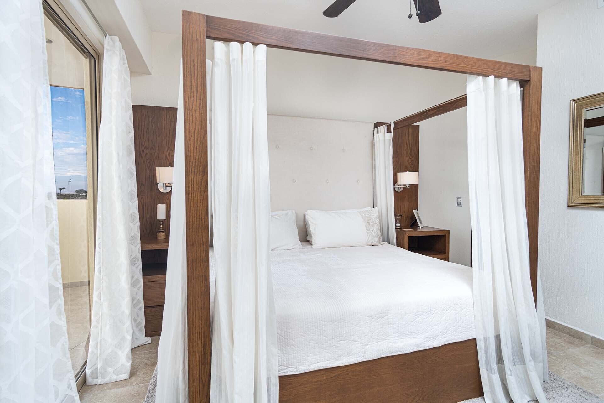 A four-poster bed with white linens, wooden headboard, and a nightstand with a lamp.