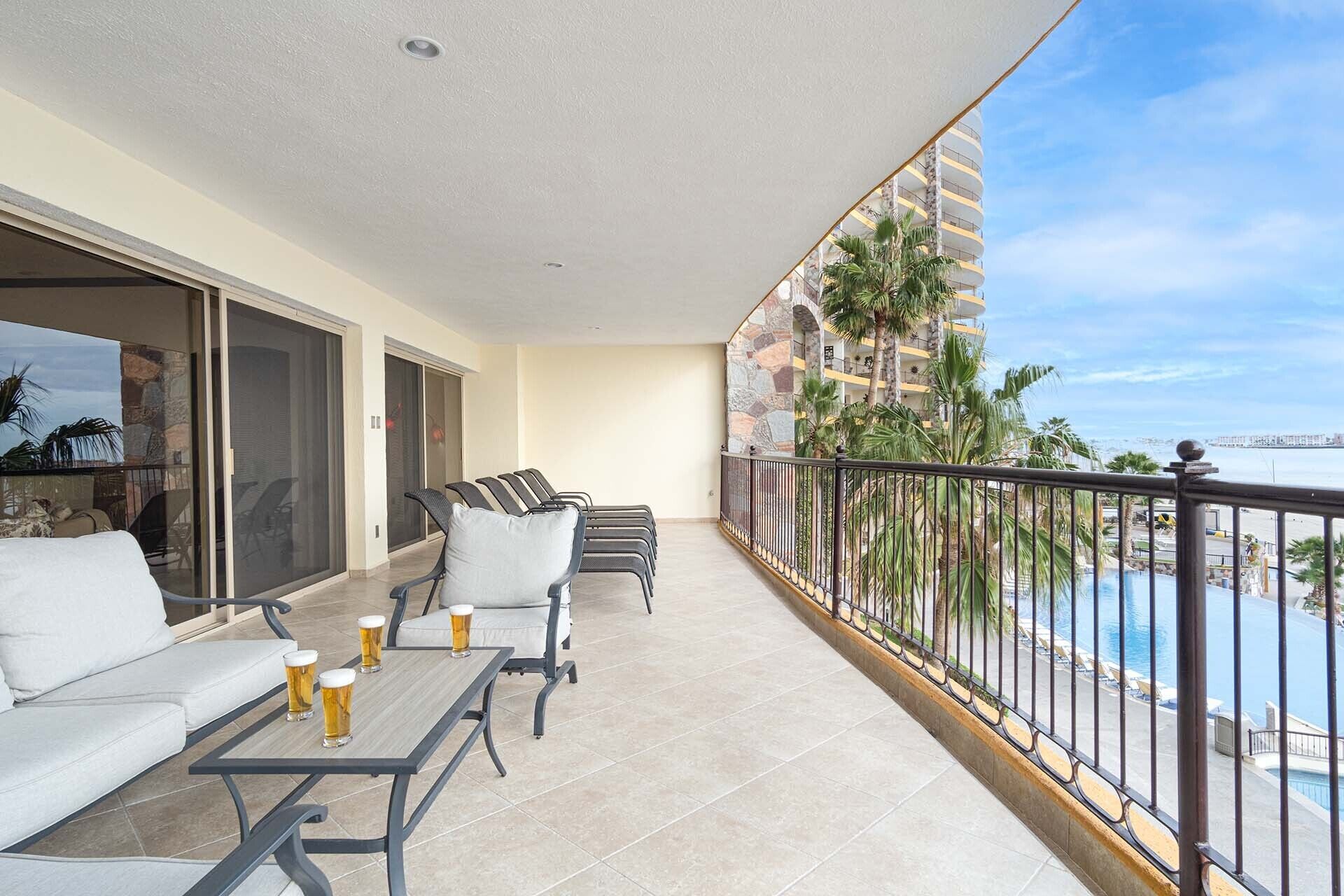 A balcony with a sofa, chairs, and a table with drinks. It overlooks a pool and a building with a stone facade.