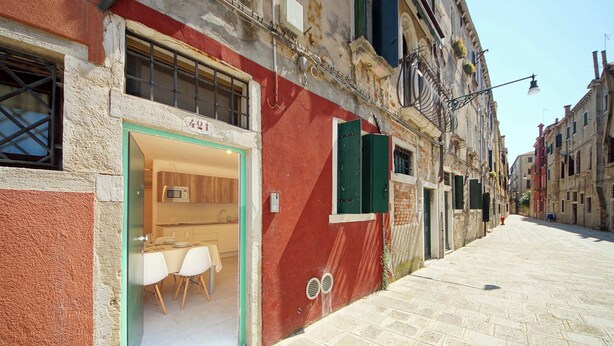 A narrow street with a building featuring a large glass door entrance, a dining area with chairs, and a red facade. - DolceVita Apartments N. 521 (Venice)