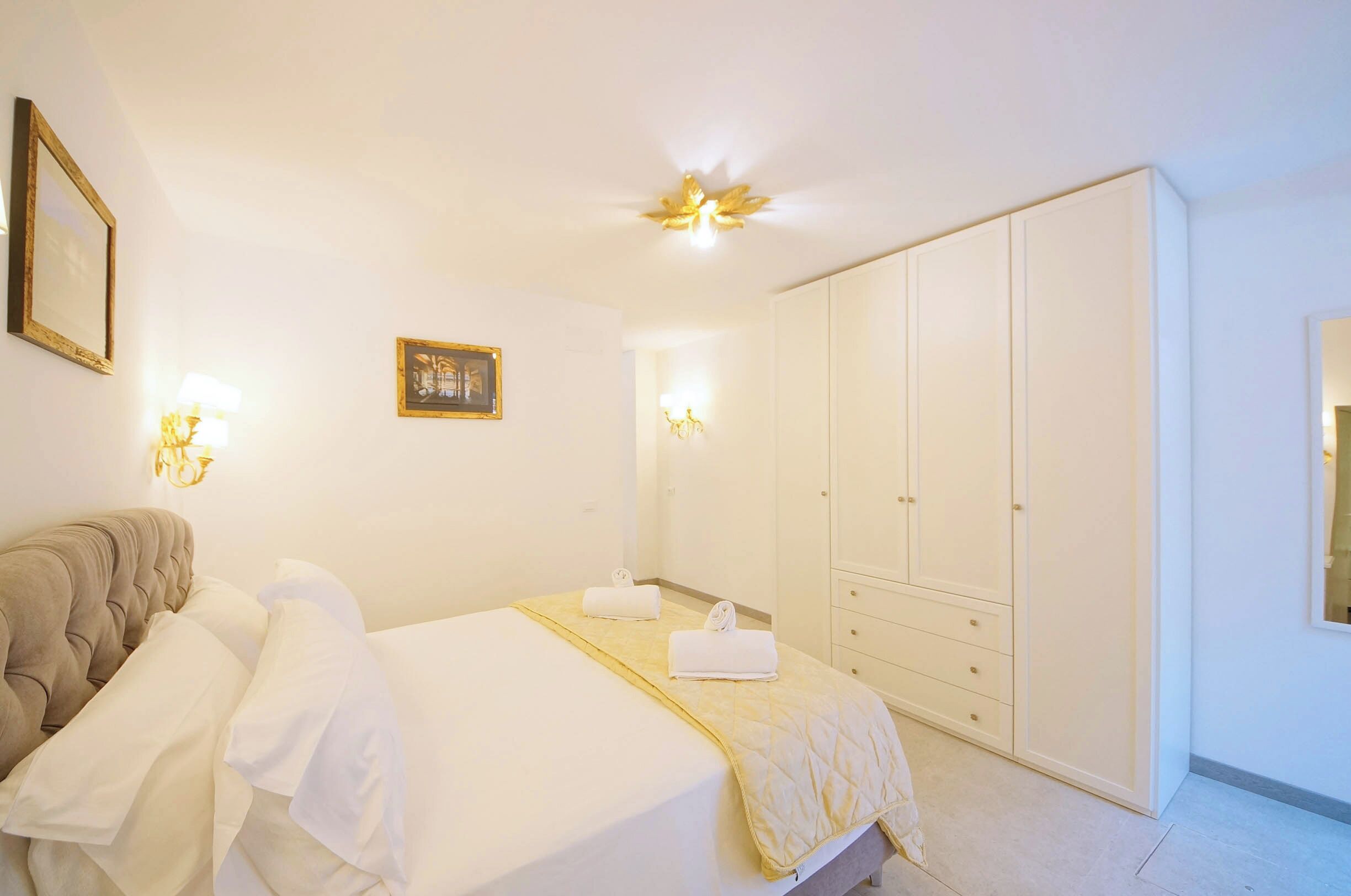A hotel room with a bed, a wardrobe, a bedside table, and wall-mounted lights.