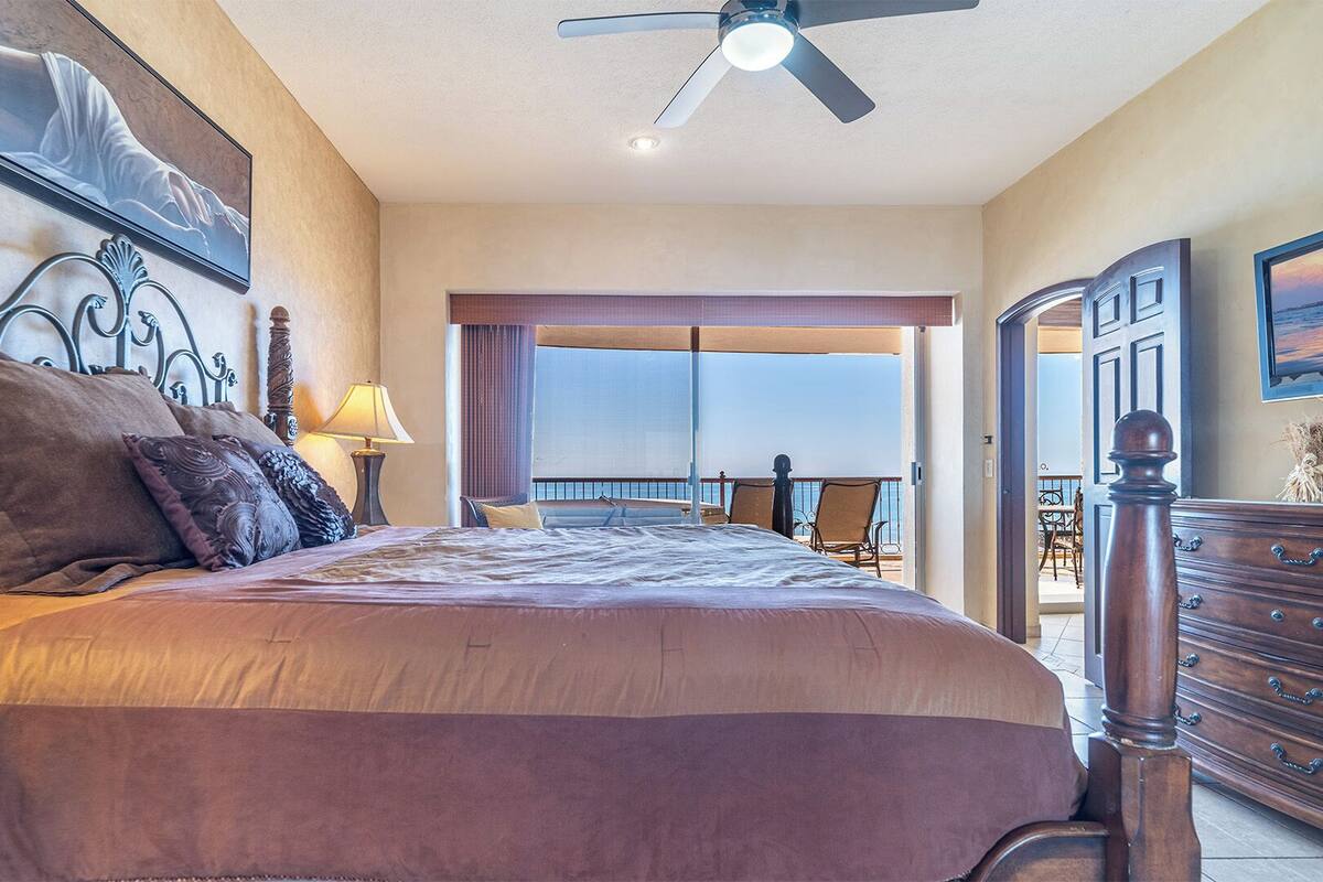 Een ruime slaapkamer met een groot bed, een plafondventilator, uitzicht op het balkon en een houten dressoir.