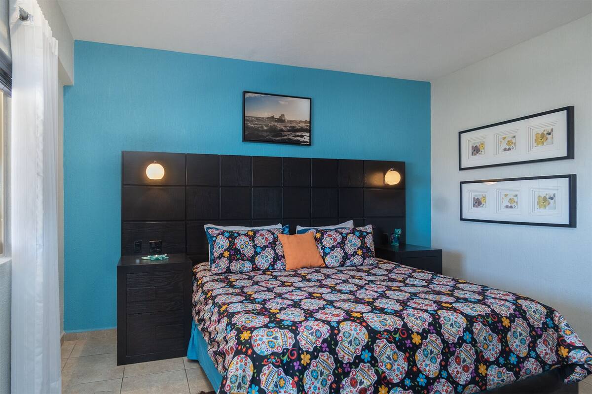 Een slaapkamer met een donker hoofdbord, een kleurrijk sprei en een ingelijst beeld boven het bed.