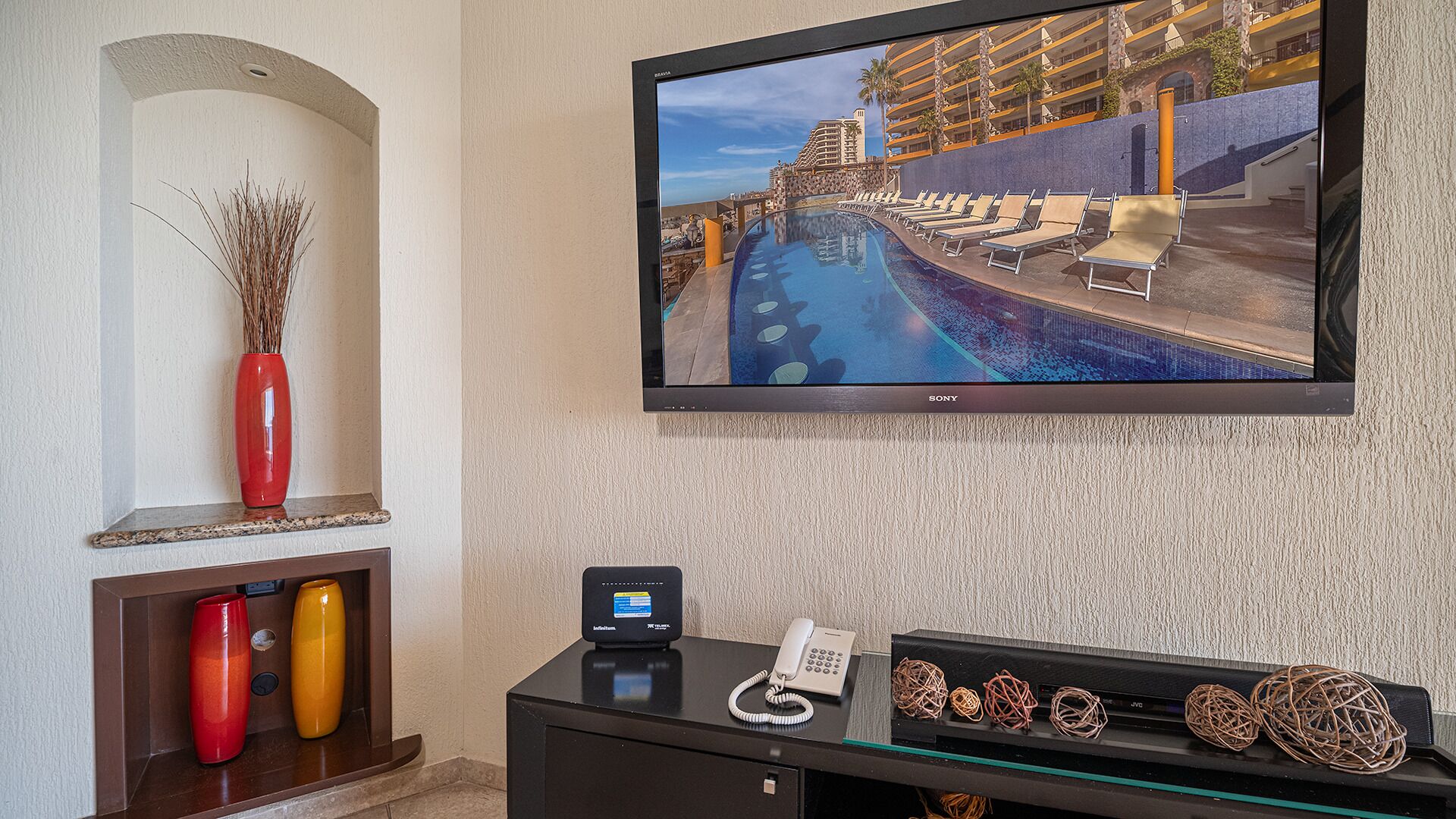 A hotel room with a large flat-screen TV mounted on the wall, a desk with a phone and a small decorative item, and a shelf with two vases.